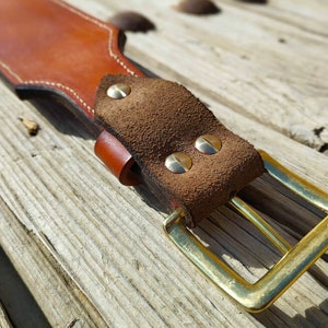 May include: A brown leather belt with a suede buckle and brass rivets. The belt is on a wooden surface.