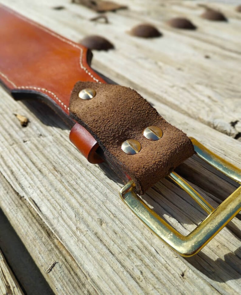May include: A brown leather belt with a suede buckle and brass rivets. The belt is on a wooden surface.