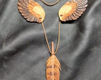 Op de afbeelding: Een handgemaakte ketting met een veervormige hanger en twee vleugelvormige elementen. De hanger en vleugels zijn gemaakt van hout, met gedetailleerd snijwerk en een natuurlijke bruine kleur. De ketting hangt aan een bruin koord.
