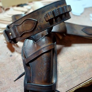 May include: A dark brown leather gun holster and matching belt set with a distressed finish. The holster features an embossed horse head design, stitched edges, and a strap. The belt includes integrated loops for ammunition storage.