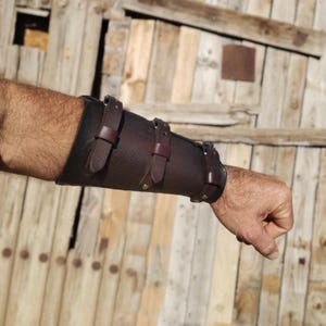 May include: A dark brown leather arm guard with three adjustable straps and silver-colored rivets. The arm guard is worn on the forearm and wrist, and is made of thick, textured leather.