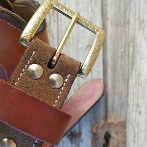 May include: Close-up of a brown leather belt with a brass buckle. The belt has a brown suede lining and two silver rivets.