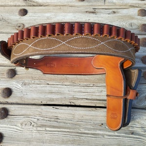 May include: A brown leather gun belt with a suede panel and a white stitching design. The belt has a cartridge loop and a holster for a pistol.