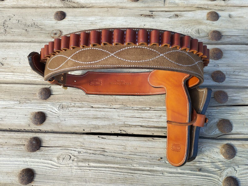 May include: A brown leather gun belt with a suede panel and a white stitching design. The belt has a cartridge loop and a holster for a pistol.