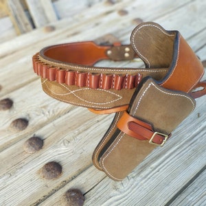 May include: A brown leather gun belt with a suede holster and a cartridge belt. The belt is decorated with white stitching.