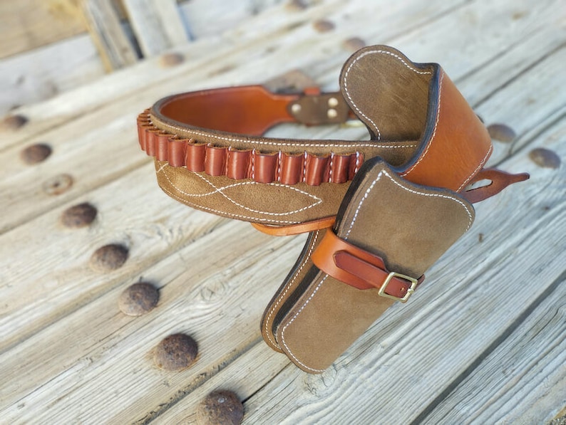 May include: A brown leather gun belt with a suede holster and a cartridge belt. The belt is decorated with white stitching.