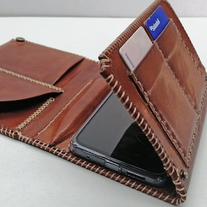 Op de afbeelding: Een bruine leren portemonnee met een telefoon en creditcards erin. De portemonnee is open en toont meerdere vakken en compartimenten. De randen zijn gestikt met een contrasterend lichtgekleurd garen. Ontworpen voor een smartphone en kaarten.