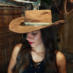 Op de afbeelding: Bruine suède cowboyhoed met een decoratieve band. De hoed heeft een brede rand en een gestructureerde kroon. De achtergrond omvat een houten muur en het woord "GUNBELTS" op een bord.