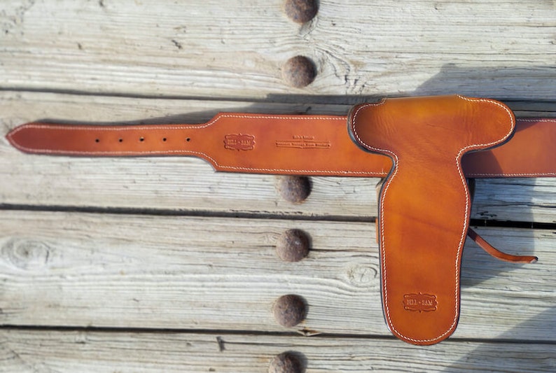 May include: A brown leather gun belt with a holster. The belt has a white stitching detail and the text "Bill-Man" is embossed on the belt and holster.