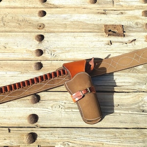 May include: A brown leather gun belt with a holster and a bullet loop. The belt has a decorative stitching pattern and a silver buckle. The holster is made of brown leather and has a flap.