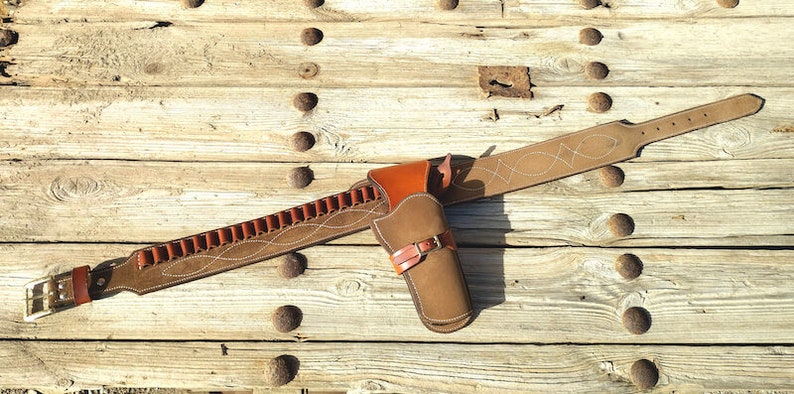 May include: A brown leather gun belt with a holster and a bullet loop. The belt has a decorative stitching pattern and a silver buckle. The holster is made of brown leather and has a flap.