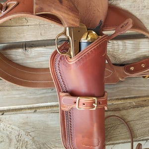 Può includere: Un revolver con impugnatura in legno e telaio in ottone inserito in una fondina in pelle marrone. La fondina presenta cuciture decorative, un cinturino con fibbia in ottone ed è fissata a un'ampia cintura in pelle con rivetti in metallo.