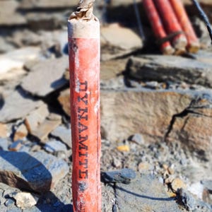 Op de afbeelding: Een rode dynamietstaaf met het woord "DYNAMITE" verticaal afgedrukt. De staaf staat op een rotsachtige ondergrond, met andere dynamietstaven op de achtergrond. Een zwarte draad loopt van de bovenkant van de staaf.