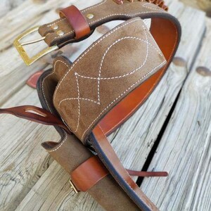May include: A brown leather gun belt with a suede holster and a cartridge belt. The belt has a brass buckle and is decorated with white stitching.