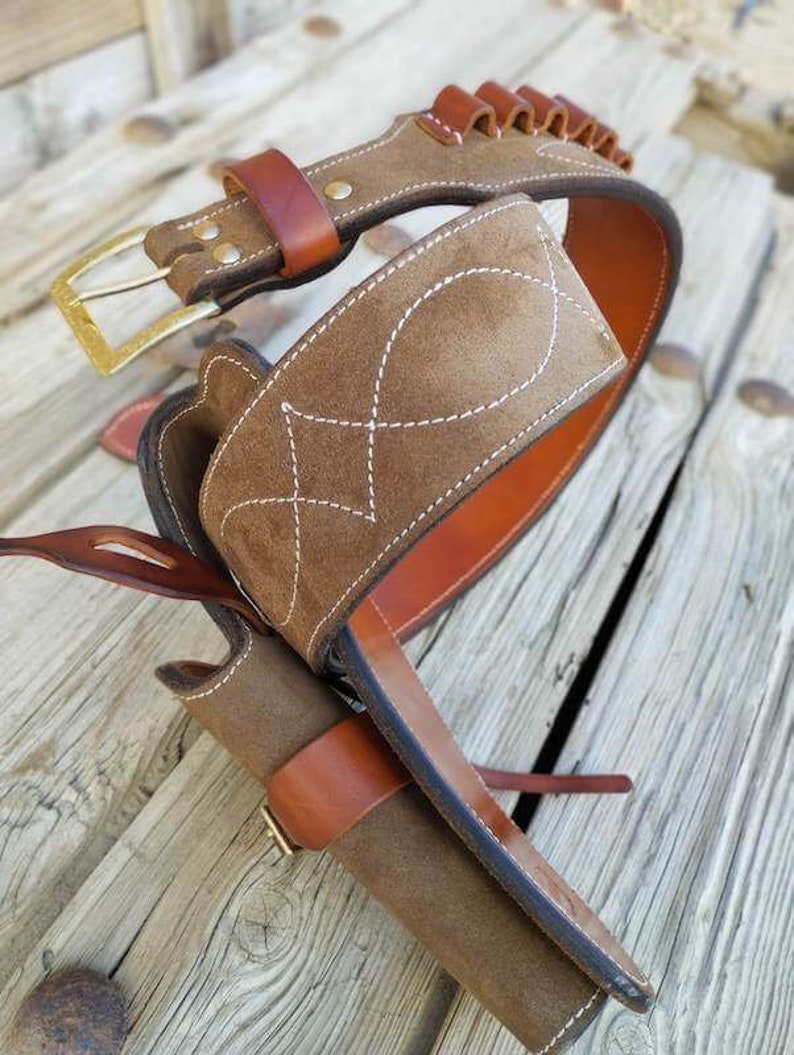 May include: A brown leather gun belt with a suede holster and a cartridge belt. The belt has a brass buckle and is decorated with white stitching.