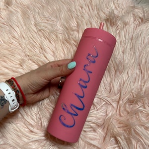 May include: A matte pink cylindrical tumbler with a matching lid and a straw. The name Chiara is written vertically on the side of the tumbler in a reflective, iridescent script font that shifts between shades of blue, purple, and green.