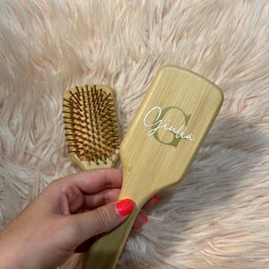Può includere: Una spazzola per capelli in legno con una finitura naturale e il nome "Giulia" inciso sul manico. La spazzola è accanto a una spazzola per capelli in legno più piccola con setole naturali.