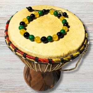 以下が含まれることがあります： 黒い、緑色、黄色のビーズがついたブレスレットが、木製のジャンベの上に置かれています。