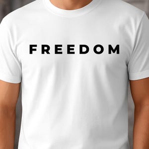 May include: White t-shirt featuring the word "FREEDOM" in bold, black capital letters. The shirt has a crew neck and short sleeves. The fabric looks to be a soft, lightweight material. A simple, minimalist design.