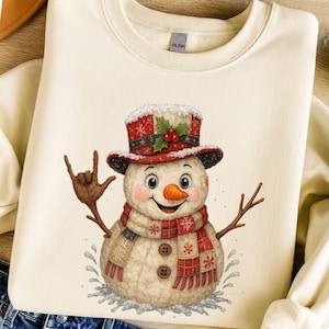 Könnte beinhalten: Cremefarbenes Sweatshirt mit einem skurrilen Schneemann-Design. Der Schneemann trägt einen rot-weißen Hut und Schal, eine Karottennase und Zweigarme. Das Design beinhaltet eine Handgeste und Schneeflocken, ideal für den Winter.