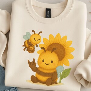 May include: Cream-colored sweatshirt featuring a cartoon bee design. The design includes two bees, a sunflower, and the sign language symbol for "I love you." One bee carries a honey pot. The sweatshirt is made of a soft material.