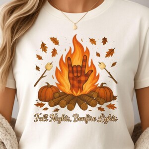 Może przedstawiać: Kremowy t-shirt z motywem ogniska. Projekt zawiera gest dłoni w kratę, płomienie, dynie i pianki na patykach. Tekst "Fall Nights, Bonfire Lights" jest wydrukowany poniżej. Odzież o tematyce jesiennej.