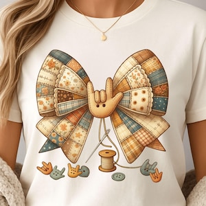 May include: Cream-colored t-shirt featuring a large, patchwork bow design with a hand making the "I love you" sign. The bow is made of various fabric patterns in shades of brown, orange, and blue. A spool of thread, needle, and buttons are also shown.