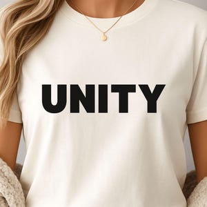 Puede incluir: Camiseta color crema con la palabra "UNITY" impresa en letras mayúsculas negras y en negrita. La camiseta tiene mangas cortas y cuello redondo. Se ve un collar de oro.