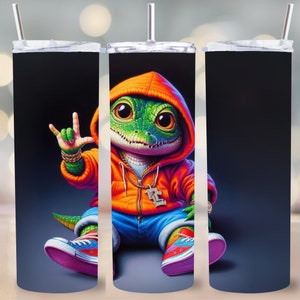 Könnte beinhalten: Ein Edelstahlbecher mit schwarzem Hintergrund und einem Cartoon-Bild eines grünen Alligators, der einen leuchtend orangen Hoodie, blaue Jeans und bunte Turnschuhe trägt. Der Alligator zeigt mit einer Hand ein Peace-Zeichen und hält in der anderen eine goldene Kette mit einem Anhänger.
