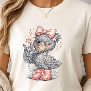 May include: Cream-colored t-shirt featuring a cartoon bird illustration. The bird is gray with a pink bow, pink boots, and is winking. The design includes hearts, stars, and water splashes. A gold necklace is visible.