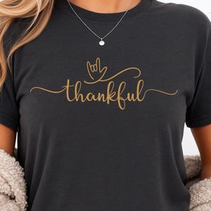 May include: Dark gray t-shirt with the word "thankful" in gold script. The word is accompanied by a hand-shaped design. The shirt is a comfortable, casual style.