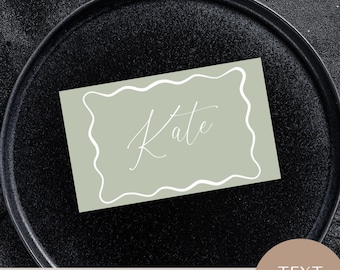 May include: Sage green place card with a white wavy border and the name "Kate" written in white cursive script. The card is on a black surface.