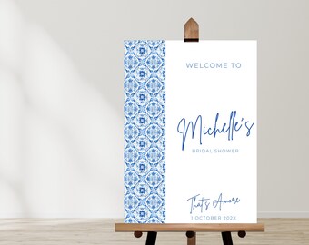 Op de afbeelding: Een wit welkomsbord met een blauwe en witte gepatroneerde rand. Het bord luidt "Welkom bij Michelle's Bridal Shower That's Amore 1 oktober 202X".