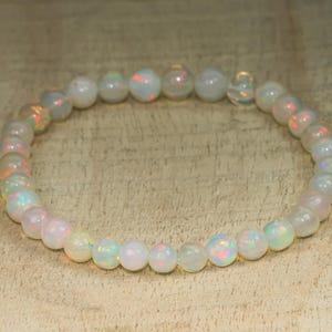 Puede incluir: Un brazalete hecho de cuentas redondas e iridiscentes de ópalo. Las cuentas son blancas con destellos de rojo, verde y naranja. El brazalete está ensartado en una banda elástica y se muestra sobre una superficie marrón claro.