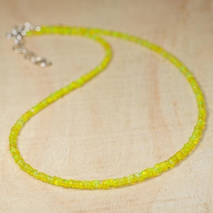 May include: A delicate necklace featuring small, round, yellow-green beads. The beads are strung together to form a necklace with a silver-colored clasp. The necklace is displayed on a light brown wooden surface.
