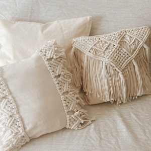 Puede incluir: Dos almohadas de color crema con ribete de flecos. Una almohada tiene un diseño de macramé tejido con un patrón geométrico.