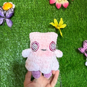 May include: A plush toy with a light pink, textured, crocheted body and two small purple feet. The toy features two prominent circular eyes with white, purple, and black spiral patterns and a small, simple horizontal black line for a mouth.