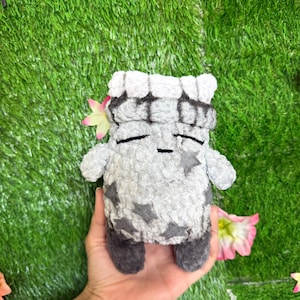 May include: A plush toy shaped like a blocky character with a light gray textured body, dark gray feet, and a dark gray top section. The character features embroidered closed eyes and a small mouth, decorated with dark gray star-shaped patches.