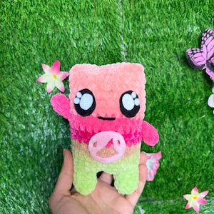 May include: A plush toy with a rectangular body featuring a gradient of peach, pink, and light green yarn. The toy has two small arms, a circular snout detail, and large black and white eyes. The texture is soft and fuzzy, resembling chenille yarn.
