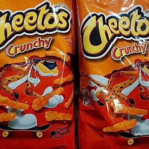 May include: Two bags of Cheetos Crunchy cheese puffs. The bags are orange and feature a cartoon cheetah wearing sunglasses and riding a skateboard. The text "Cheetos Crunchy" is printed on the front of the bags.