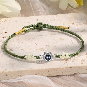 May include: A green string bracelet with white flower charms and a silver charm with a black pearl in the center.