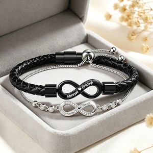 May include: Two infinity symbol bracelets in a grey gift box. One bracelet is black braided leather with a black infinity charm engraved with "Mireyal Erica!". The other is silver with a diamond-studded infinity charm engraved with "Smith David".