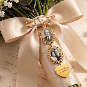 Pode incluir: Um broche dourado com duas molduras ovais para fotos e um pingente em forma de coração gravado com o texto "You are always in my heart", preso a uma fita de cetim larga na cor champanhe pálido em torno de um buquê de rosas brancas.