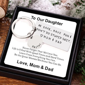 Puede incluir: Un llavero plateado con un dije en forma de corazón y una etiqueta que dice "To Our Daughter Be Safe. Have Fun & Don't Do Stupid Shit. ♡ Mom & Dad" y una etiqueta más pequeña que dice "Skylar" con el texto "Life is Filled With Good Times and Hard Times. Never Forget That We Love You. Learn From Everything You Can. Be the Great Woman We Know You Can Be. Love, Mom & Dad".