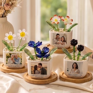 Puede incluir: Cuatro macetas cuadradas de cerámica blanca sobre bases redondas de madera, cada una con flores de vidrio y una foto personalizada. Las macetas incluyen textos como "To Our Special Grandparents", "Our Love Grows Still", "To My Soulmate and Partner" y "Happy First Father's Day".