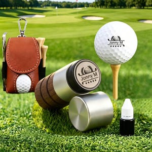 May include: A brown leather golf ball pouch with a metal clip, a white golf ball on a wooden tee, a cylindrical wooden and silver golf ball stamper with the text Jonny M, and a small black ink bottle for marking golf balls.