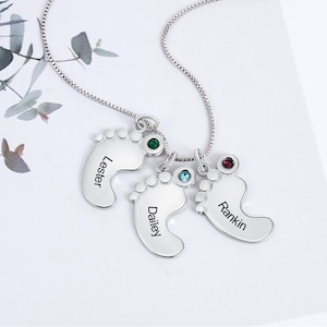 May include: A silver necklace with three baby footprint pendants. Each footprint charm has a different coloured gemstone and a name engraved: Lester, Dailey, and Rankin. The necklace is displayed on a white surface.