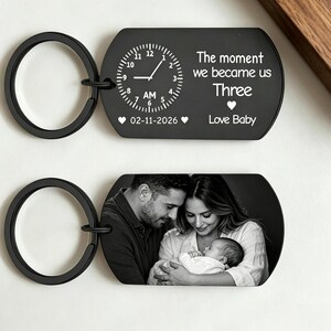 May include: Two black keychains. One features a clock design with the text "The moment we became us Three Love Baby" and the date 02-11-2026. The other keychain displays a black and white photo of a family.