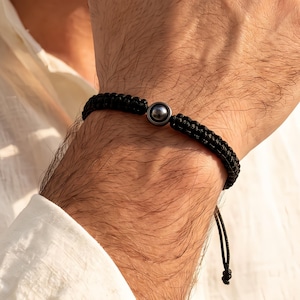 Könnte beinhalten: Ein schwarzes Armband aus geflochtener Kordel mit einer einzelnen, mittig platzierten metallischen Kugelperle. Das Armband ist geflochten und verfügt über verstellbare Zugbänder am Ende, die einen sicheren Sitz am Handgelenk ermöglichen.