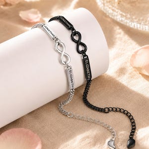 Op de afbeelding: Twee kettingarmbanden met oneindigheidssymbolen en rechthoekige metalen staafjes gegraveerd met de namen JONATHAN en ELIZABETH. De ene armband is zilverkleurig en de andere is zwart. Elke ketting eindigt met een kleine halve hartbedel.